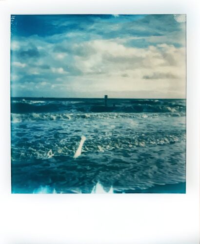 Colour Polaroid photo of breaking waves on the shore with a groyne post out in the sea and a blue and cloudy sky behind